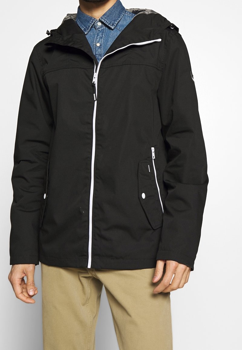 Homme portant une veste à capuche noire avec une fermeture éclair blanche et un pantalon kaki, debout devant un fond blanc.