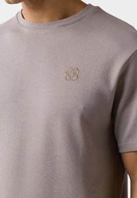 Άνδρας που φοράει μωβ βαμβακερό κοντομάνικο μπλουζάκι με μικρό κεντημένο μπεζ λογότυπο στο αριστερό στήθος.