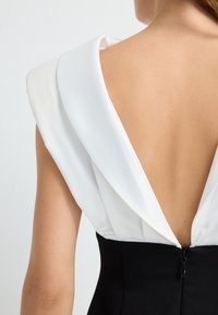 Zwarte jurk met een witte geplooide kraag en een diepe V-hals aan de achterkant, met een zichtbare ritssluiting. Gladde textuur met contrasterende kleuren.