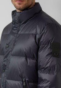 Duvetirana crna jakna s visokim ovratnikom, s kopčanjem na gumbe, bočnim patentnim zatvaračima i teksturiranim tkivom na rukavu.