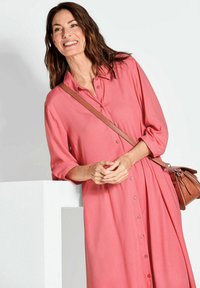 Langes, pinkes Hemdkleid mit Dreiviertelärmeln und lässigem Schnitt. Hält eine braune Umhängetasche mit einem schlichten Design.