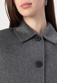 Manteau en laine grise avec un col, deux boutons noirs et une texture feutrée. Gros plan mettant en évidence le matériau et le détail des boutons.