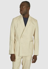 Beige linnen dubbelzijdig colbert met twee voorkeurszakken en lichte knopen. Getextureerde stof, getailleerde pasvorm, en met nietjes revers.