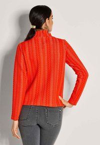 Roter strukturierter Strickpullover mit vertikalen Streifen, langärmelig und mit hohem Kragen, kombiniert mit dunkelgrauen Jeans.