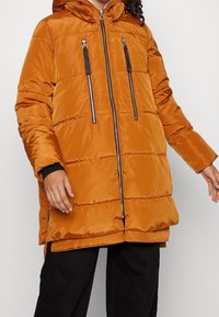 Oranssi puhvitakki hupulla, jossa on mustat vetskarit ja kaksi etutaskua, kiiltävän, tikatun pinnan kanssa.