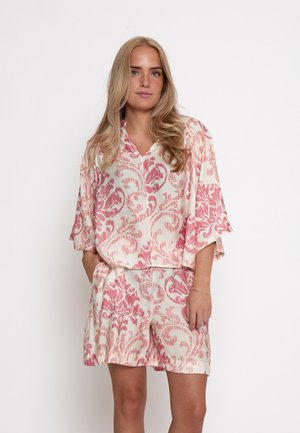 Femme blonde debout, portant une tenue ample crème avec un motif botanique rose, un haut à manches courtes et un short assorti, main dans la poche.