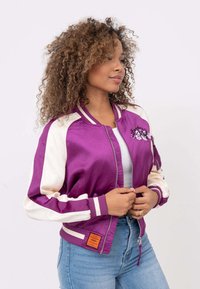 Fioletowa satynowa kurtka bomberka z białymi rękawami, ściągaczami oraz pomarańczową metką. Posiada kwiatowy haft na lewej piersi.