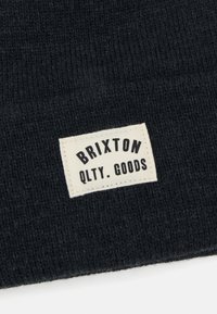 Fekete kötött sapka, puha tapintással, téglalap alakú krémszínű címkével, amelyen a "BRIXTON QLTY. GOODS" felirat fekete betűkkel olvasható.