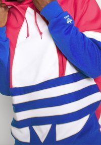 Färgglad hoodie med en blandning av röd, blå och vit. Den har en stor triangulär design och flera horisontella ränder med en texturerad finish.