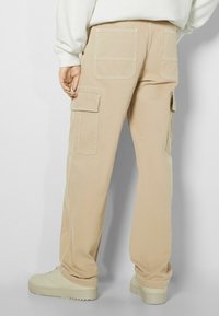 Beige Cargo-Hosen mit mehreren Taschen, sichtbaren Nähten, geraden Beinen und einem leichten Glanz im Stoff. Kombiniert mit hellbeigen Sneakers.