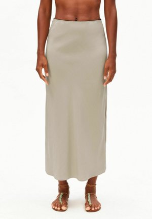 Frau trägt einen hoch taillierten, beigefarbenen Rock in Knöchellänge und braune Sandalen mit grünen Perlenakzenten, steht vor einem weißen Hintergrund.