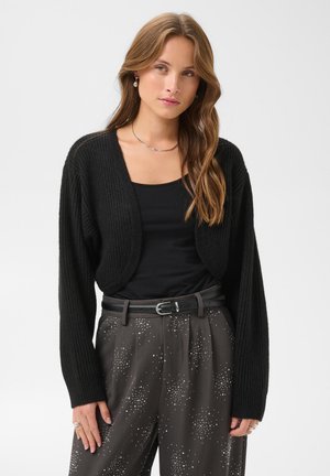 Femme portant un cardigan court noir par-dessus un top noir et un pantalon sombre taille haute avec un motif à pois blancs et une ceinture noire.