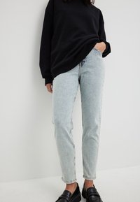 Jean en denim bleu clair avec une coupe fuselée, texture déchirée et détails brodés, associés à un sweat-shirt noir surdimensionné et des mocassins noirs.
