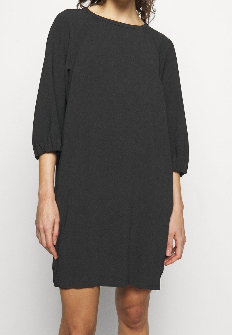 Femme porte une robe noire ample avec des manches trois-quarts resserrées aux poignets, debout devant un fond clair uni.