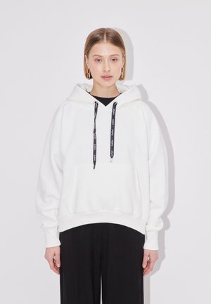 Ung kvinde med kort hår iført en hvid hoodie med sorte snører og sorte bukser, står mod en hvid baggrund.
