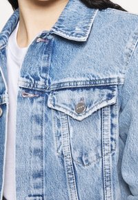 Ljusblå denimjacka med två bröstfickor, synlig sömnad och slitna detaljer på tyget. Knappdetaljer på fickorna.