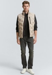 Beige quiltad vadderad väst med en ficka med dragkedja, bärs över en svart långärmad tröja, kombinerad med mörka jeans och vita sneakers.