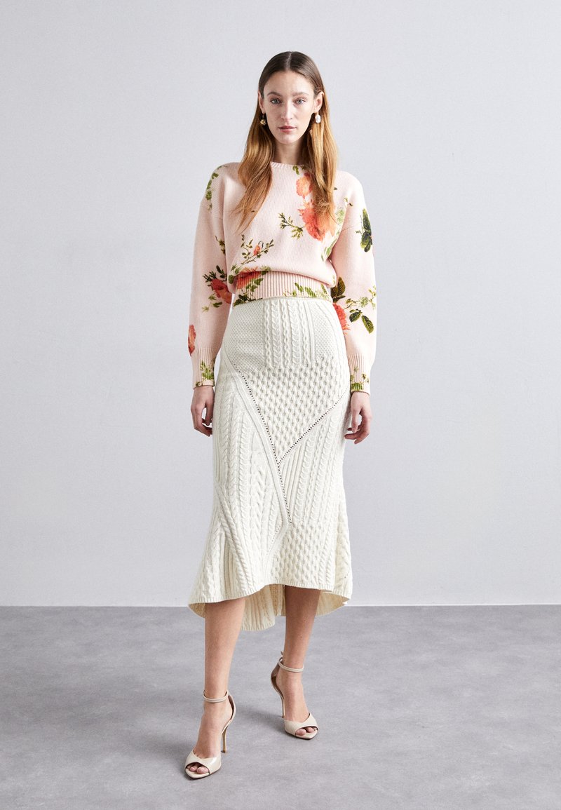 Pull rose à fleurs avec des manches longues, associé à une jupe midi en tricot blanche avec des motifs texturés et un ourlet asymétrique. Talons beiges.