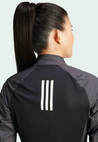 Czarna kurtka sportowa z szarymi akcentami, z wysokim kołnierzem i trzema białymi pionowymi paskami na plecach, z teksturowanym wzorem materiału.