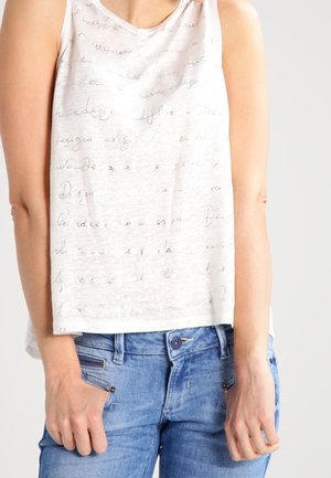 Ärmelloses weißes Oberteil mit schwach sichtbarem, kursiv geschriebenem Text, getragen von einer Person in blauer Jeans, die mit den Händen nahe den Taschen steht.