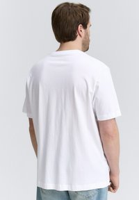 Camiseta blanca de algodón con mangas cortas, cuello redondo y textura suave, mostrada desde la parte posterior con un fondo neutro.