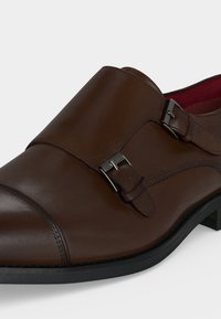 Chaussure habillée en cuir marron à double bride de moine, boucles argentées et semelle noire, vue rapprochée de côté.