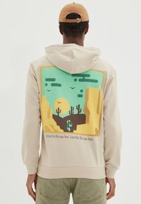 Felpa con cappuccio beige con una stampa grafica di una scena desertica, caratterizzata da cactus verdi, un sole giallo e montagne stilizzate sul retro.