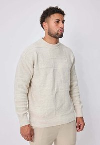 Homme aux cheveux courts et bouclés, portant une pull en tricot texturé beige clair et un pantalon beige, se tenant devant un fond blanc uni.