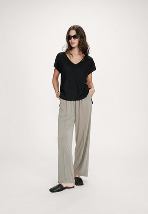 Femme portant un haut noir à manches courtes en V, pantalon gris à jambes larges, chaussures noires sans lacets et lunettes de soleil, debout les mains dans les poches.