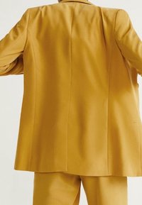 Costume jaune avec un blazer structuré et un pantalon assorti. Les caractéristiques clés incluent un tissu lisse, une coupe ajustée et des lignes épurées.