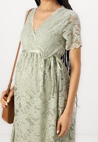 Pitsinen kietaisumekko pehmeässä vihreässä, jossa on lyhyet hihat, v-kaula-aukko ja kukkakuvio. Mekossa on satiininauhavyö vyötäröllä ja aaltoilevat pitsireunat.