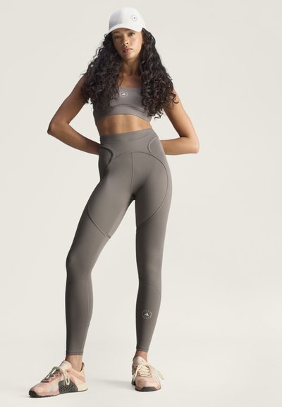 Athletic woman with long curly hair wearing gray sports bra, matching high-waisted leggings, white cap, and pink sneakers standing with hands on hips.