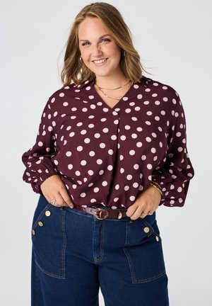 Lachende vrouw met schouderlang haar, die een kastanjebruwe blouse met roze stippen en een hoge taille blauwe spijkerbroek met gouden knopen draagt.