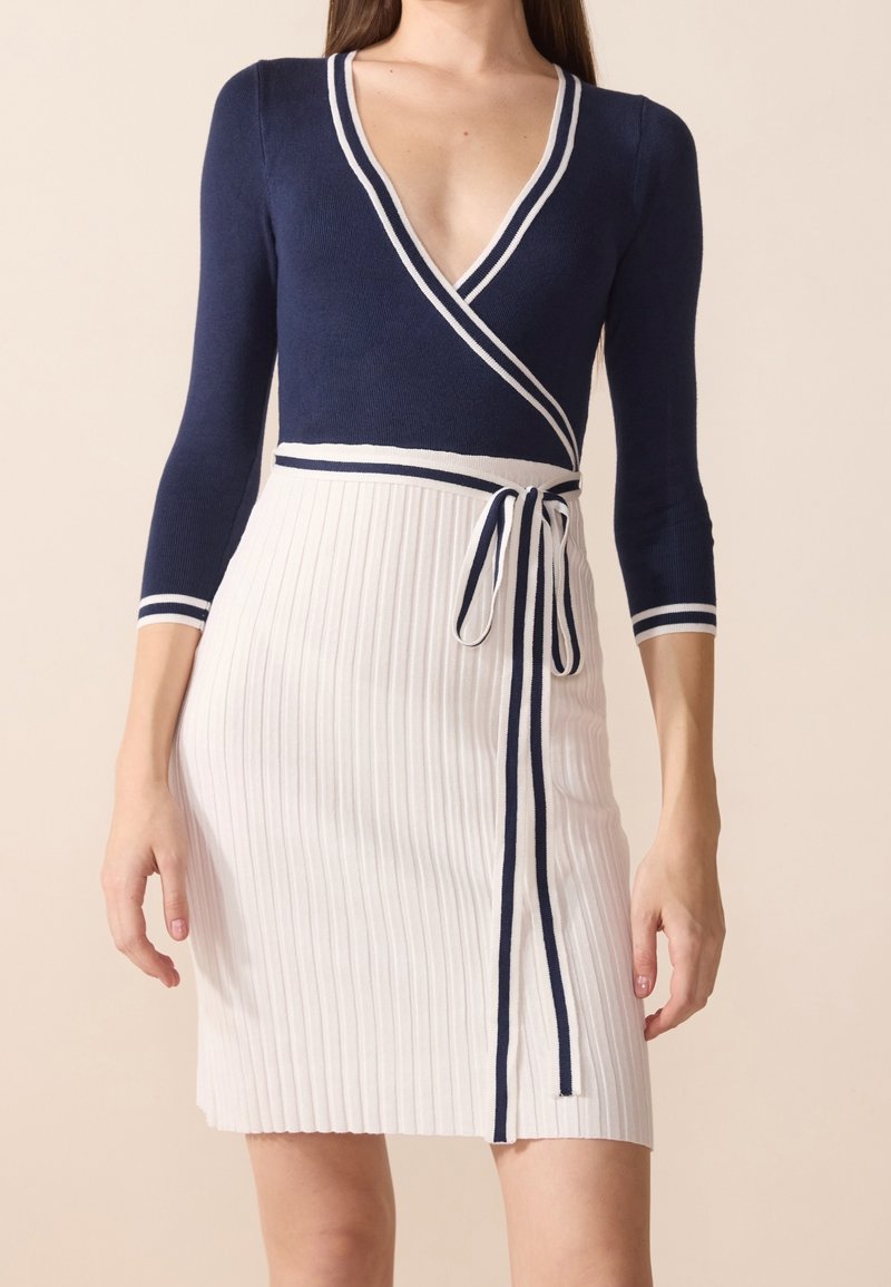 Haut bleu marine style cache-cœur avec bordure blanche, associé à une jupe blanche côtelée avec une ceinture rayée bleu marine attachée à la taille.