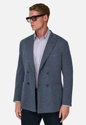 Homme portant un blazer croisé gris foncé, une chemise à rayures claires, un pantalon foncé et des lunettes de soleil noires, regardant sur le côté sur un fond blanc.