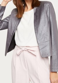 Blazer gris métallisé à coupe ajustée, ouvert sur le devant sans boutons, associé à un haut blanc et un pantalon taille haute rose pâle.