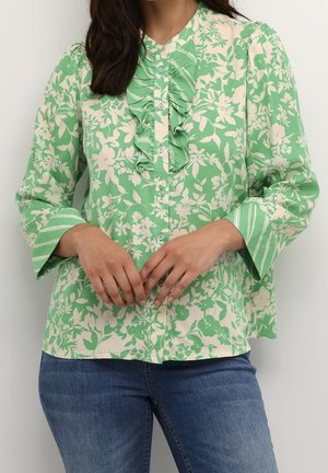 Femme portant un chemisier long à manches longues fleuri vert et crème avec un devant à volants et des poignets rayés, associé à un jean en denim bleu.