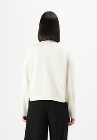 Persona con capelli neri lisci indossa una maglietta bianca a maniche lunghe e pantaloni neri, in piedi di spalle contro uno sfondo bianco uniforme.