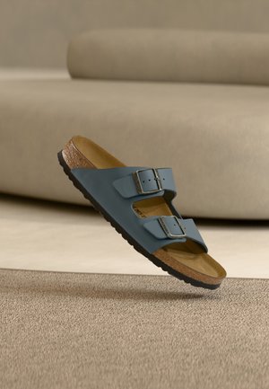 Sandale bleue à deux brides avec boucles ajustables en argent, semelle intérieure en liège et semelle noire, positionnée au-dessus d'un tapis beige texturé.