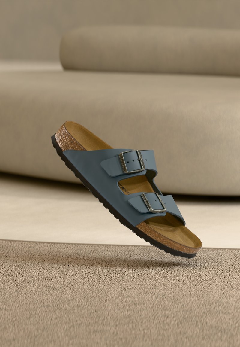 Μπλε σανδάλι με δύο λουράκια και ρυθμιζόμενες ασημί αγκράφες, πάτο από φελλό και μαύρη σόλα, τοποθετημένο πάνω σε μπεζ υφασμάτινο χαλί.