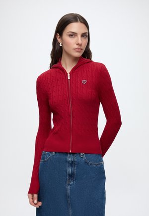 Femme aux longs cheveux foncés porte un pull rouge à torsades zippé avec un patch en forme de cœur et une jupe en jean bleue sur un fond uni.