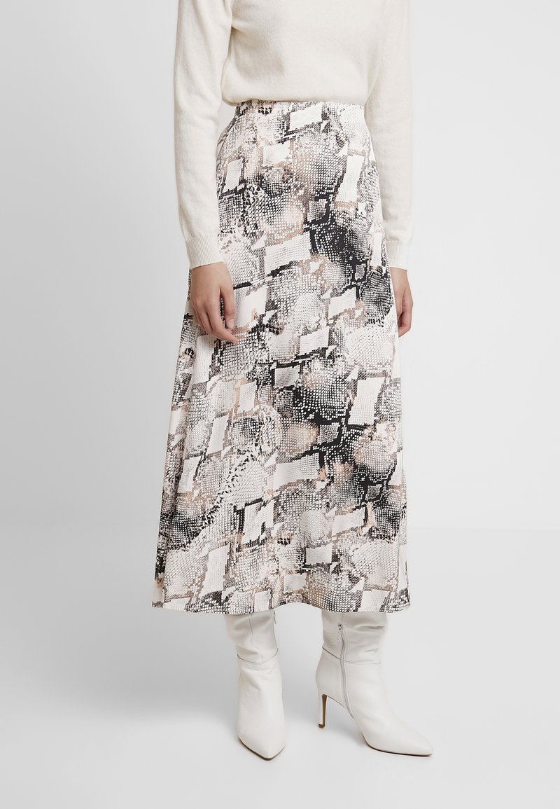 Femme portant une jupe taille haute, longueur cheville, avec un imprimé serpent noir et beige, un pull blanc et des bottes blanches à talons hauts.