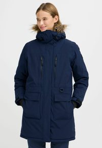 Mornarsko modra parka jakna s kapuco obloženo z naravnim krznom, več žepov z zadrgo in nalepkami ter eleganten dizajn. Vodoodporna, toplotno izolirana in trpežna tkanina.