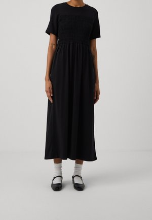 Schwarzes knielanges Kleid mit einem gerafften Oberteil, kurzen Ärmeln und einem lockeren, fließenden Rock. Getragen mit weißen Socken und schwarzen Schuhen.