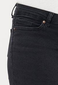 Črne denim hlače z gladko teksturo, ki imajo pet žepov, šivane detajle in majhen gumb v barvi bakra.