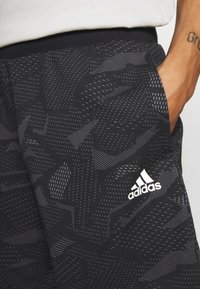 Svarta Adidas träningsshorts med abstrakta grå mönster och en vit logotyp, bärs av en person med en tatuerad arm och handen i fickan.