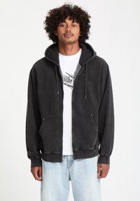 Sudadera con cremallera negra de textura suave, que presenta una capucha con cordón, bolsillos frontales y un logo bordado. Llevada sobre una camiseta gráfica blanca.