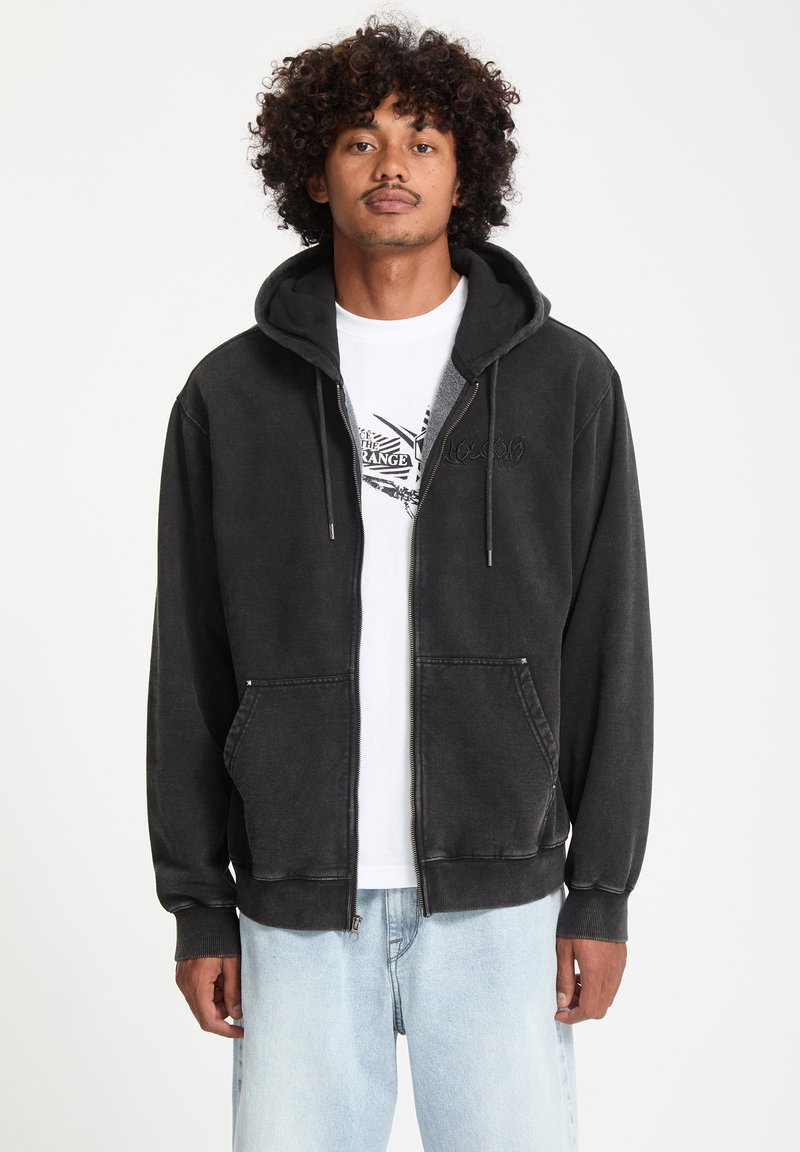 Veste à zip noire avec une texture douce, dotée d'une capuche à cordon, de poches avant et d'un logo brodé. Portée par-dessus un t-shirt graphique blanc.