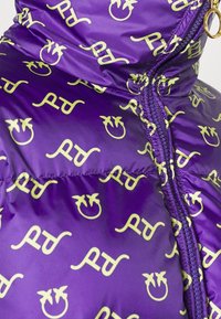 Paarse pufferjas met een herhaald geel grafisch patroon met een glimlachend gezicht en gestileerde tekst, met zichtbare ritsen en stiksels.