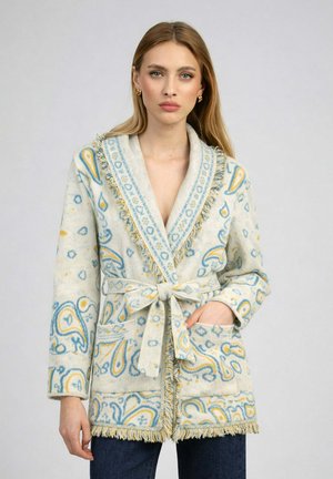 Femme portant un cardigan crème à motifs avec des dessins paisley bleus et jaunes, des bords frangés et une ceinture nouée, debout devant un fond uni.